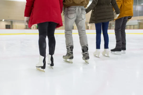 Four individuals enjoying ice skating indoors, seen from the back, all clad in winter coats and equipped with skates at the rink, wearing products by Lorem Ipsum.