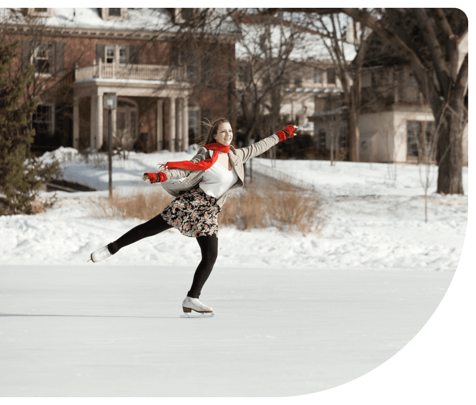 A person ice skating outdoors with arms outstretched, wearing a red scarf and a floral skirt. Snow-covered ground and buildings in the background.